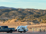 An RV rental: a white teardrop trailer is hitched to a gray pickup truck at a desert campsite, with mountains in the sunny background.