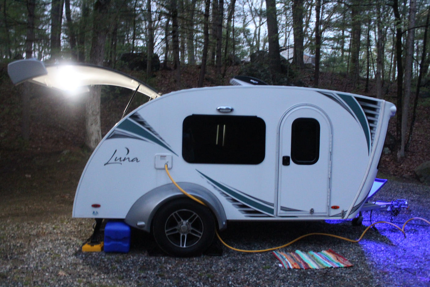 A cozy Luna teardrop camper, illuminated by bright white and blue lights, set up at a wooded campsite. Perfect for an rv rental.