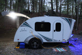 A cozy Luna teardrop camper, illuminated by bright white and blue lights, set up at a wooded campsite. Perfect for an rv rental.