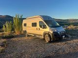 This silver camper van rv rental is parked in golden light, with tall plants & distant mountains under a blue sky.