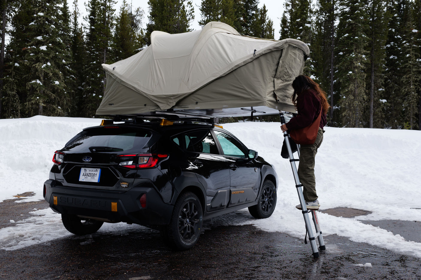 Person setting up Thule roof-top tent on black Subaru Crosstrek amidst snowy trees, ideal for an RV rental alternative.