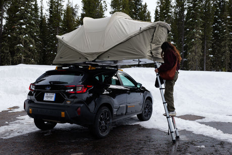 Person setting up Thule roof-top tent on black Subaru Crosstrek amidst snowy trees, ideal for an RV rental alternative.
