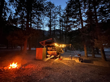 Cozy night camping. A bright campfire illuminates an RV rental trailer with a rooftop tent in a forested setting.