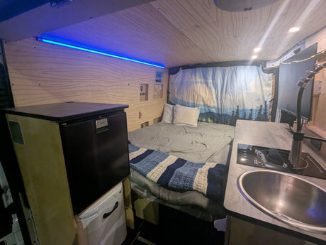 This RV rental interior features a comfortable bed, compact kitchen with sink and cooktop, and cool blue accent lighting.