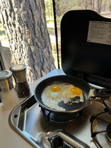 Frying eggs on a Suburban stove in an RV rental, with salt/pepper shakers. Tree and nature visible outside the window.
