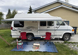 White RV rental with awning extended. Camping chairs, table, and firewood on a blue outdoor mat, ready for use.