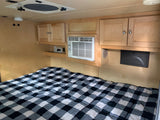 Experience this cozy RV rental bedroom. It features a bed with plaid bedding, overhead cabinets, and a convenient AC unit.