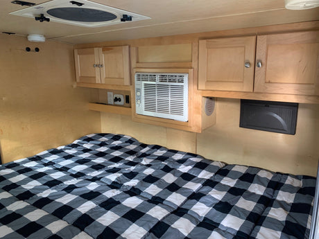 Experience this cozy RV rental bedroom. It features a bed with plaid bedding, overhead cabinets, and a convenient AC unit.