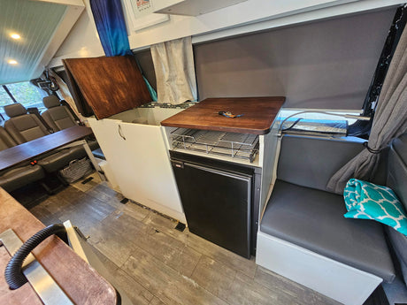 RV rental interior: kitchenette with sink/fridge, dinette table, and comfy bench seating on wood-look floor. Travel ready.