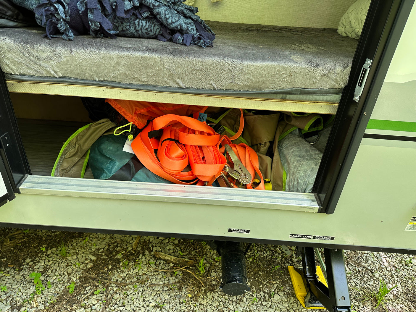 RV rental under-bed storage filled with camping gear. Exterior reveals wastewater tanks and a stabilizer jack.