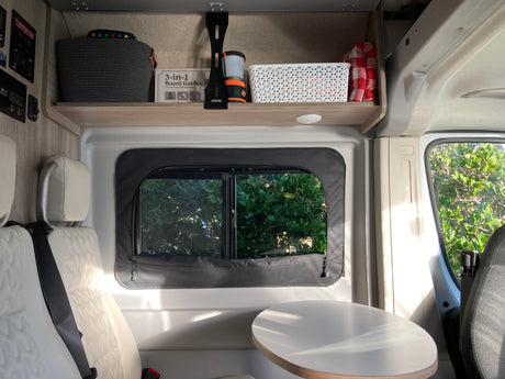 Inside an RV rental: compact living area with comfy seats, a table, shaded window, and overhead storage for games & essentials.