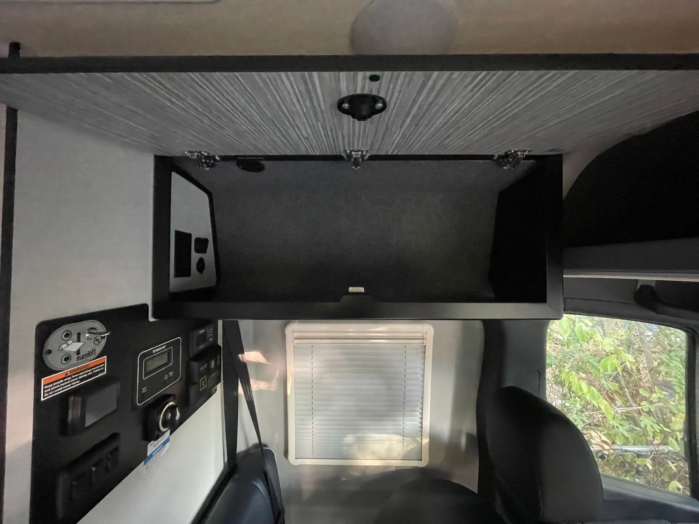 RV rental interior: Open overhead storage, a comprehensive control panel on the left, and a passenger window with blinds.