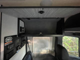 RV rental interior: Open overhead storage, a comprehensive control panel on the left, and a passenger window with blinds.