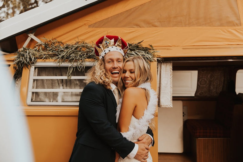 A joyful couple, the man in a crown, hugs before an orange camper van RV rental, decorated with greenery. — photo 13