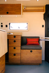This rv rental camper van boasts warm wood cabinetry, efficient storage, a cozy bench seat with a patterned pillow, and a bright window.