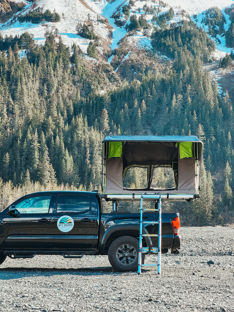 Black pickup with rooftop tent, an RV rental option, on a gravel bed with snowy mountains & pines in the background. — photo 5