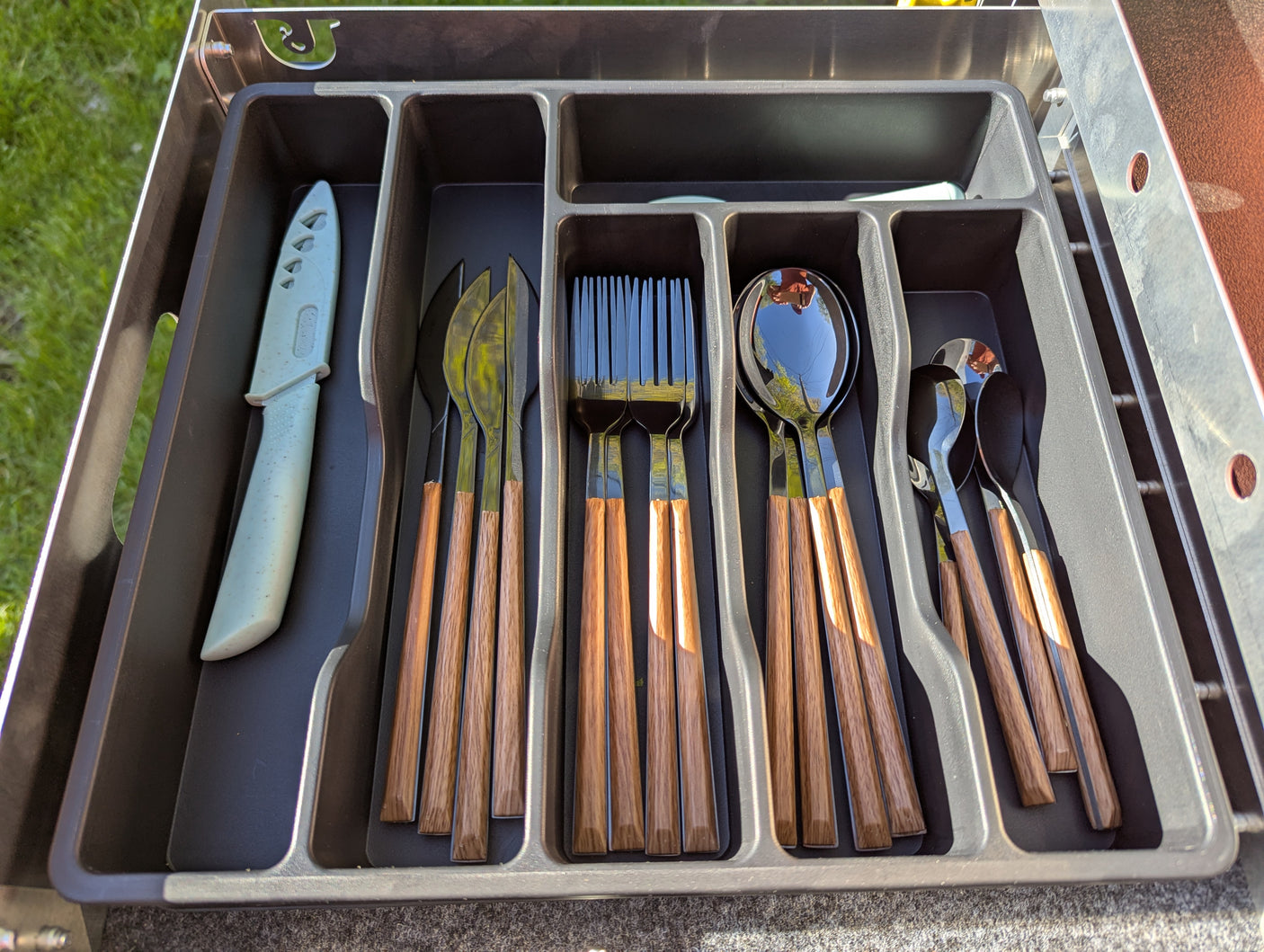 An RV rental's organized cutlery drawer: knives, forks, and spoons with wood handles and a green knife in a tray.