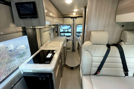 Inside an rv rental: a light-toned interior with a kitchen (sink, stovetop, fridge), TV, and passenger seating.