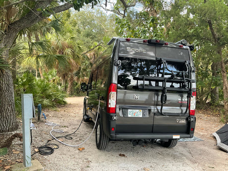 This RV rental camper van is parked at a shaded campsite, connected to electrical and water hookups, surrounded by lush vegetation.