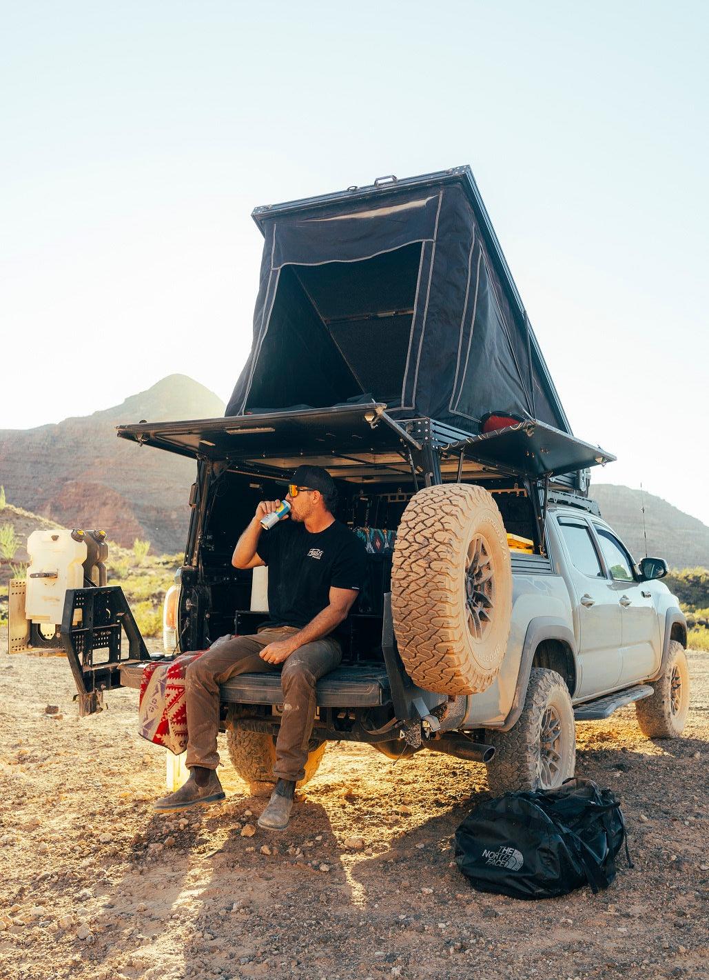Overland RV rental adventure: A man relaxes on his pickup truck's tailgate, with a pop-up camper, in a desert setting.