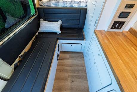Inside an RV rental: dark L-shaped seating, wood-look floor, light cabinets, and a wooden counter with power outlets.