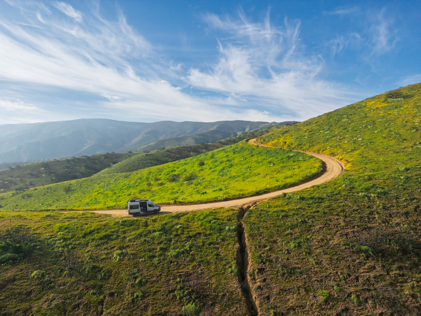 An rv rental is parked on a winding dirt road up vibrant green hills dotted with yellow wildflowers under a blue sky. Remote scenic travel.