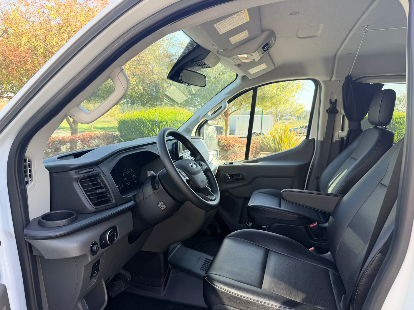 Clean front cabin of an RV rental van, featuring black seats, steering wheel, and dashboard. Bright view outside.