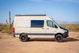 Tan camper van RV rental in a desert landscape with saguaro cacti and mountains under a clear blue sky.