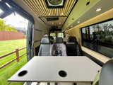 Modern RV rental van interior: spacious table with cupholders, rear passenger seats (one with a car seat), and custom wood ceiling.