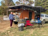 Woman smiles next to her orange tear-drop camper with an awning setup, perfect for an RV rental getaway in nature.