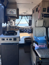 Explore this RV rental: compact kitchen, cozy bed with a scenic lake view, and a small lounge area inside.
