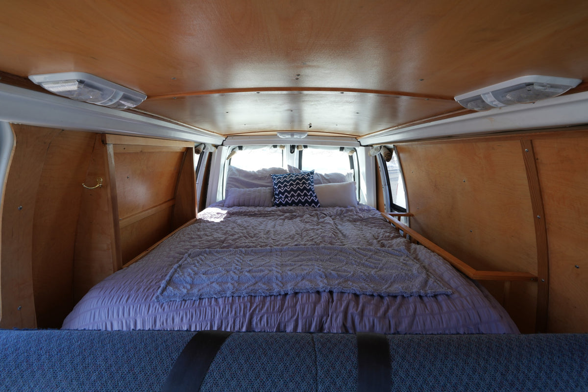 This cozy RV rental offers a bed inside a wood-paneled van, featuring ceiling lights and natural light from rear windows. — photo 10