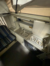 Compact RV rental interior: kitchen with sink, two-burner stove, fridge, and seating area. Features a pop-up roof.