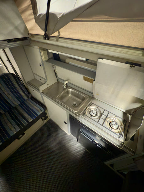 Compact RV rental interior: kitchen with sink, two-burner stove, fridge, and seating area. Features a pop-up roof.