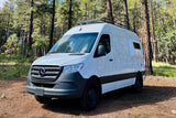 White Mercedes-Benz Sprinter RV rental parked on a dirt path in a sunny pine forest, ready for your next adventure.