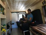 A man smiles in a cozy, wood-paneled camper van interior with custom shelves & bed. Great for an rv rental & mobile living.
