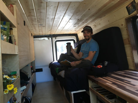 A man smiles in a cozy, wood-paneled camper van interior with custom shelves & bed. Great for an rv rental & mobile living.