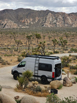 White VANSPEED RV rental parked on a sandy path amidst Joshua trees, large boulders, and desert mountains.