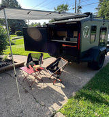 An RV rental: a dark green travel trailer with its back hatch open to reveal a kitchen. An awning extends, with two chairs & a table set up.
