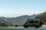 Embrace adventure! This RV rental offers stunning mountain views. A person enjoys van life freedom, standing on the van's roof.