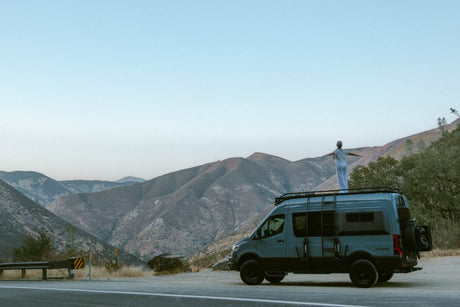 Embrace adventure! This RV rental offers stunning mountain views. A person enjoys van life freedom, standing on the van's roof.