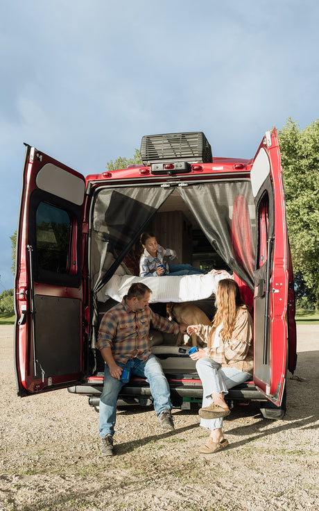 Inside an **rv rental**: A family and dog relax in their red camper van with open doors, enjoying the outdoors.