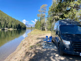 Serene RV rental campsite: Dark van by a calm river, surrounded by forests, with distant mountains and blue sky. Chairs and rug invite relaxation.