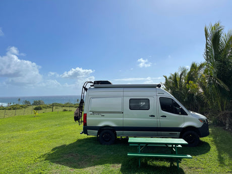 Silver Winnebago Revel 4x4 rv rental on green lawn, overlooking ocean, with palm trees & picnic table.
