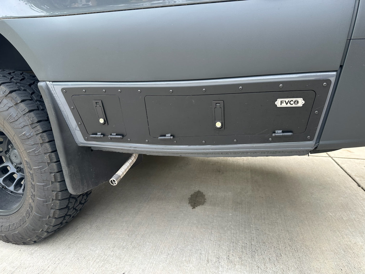 Modified RV rental van side with custom storage compartments and exhaust. Shows an off-road tire and concrete stain.