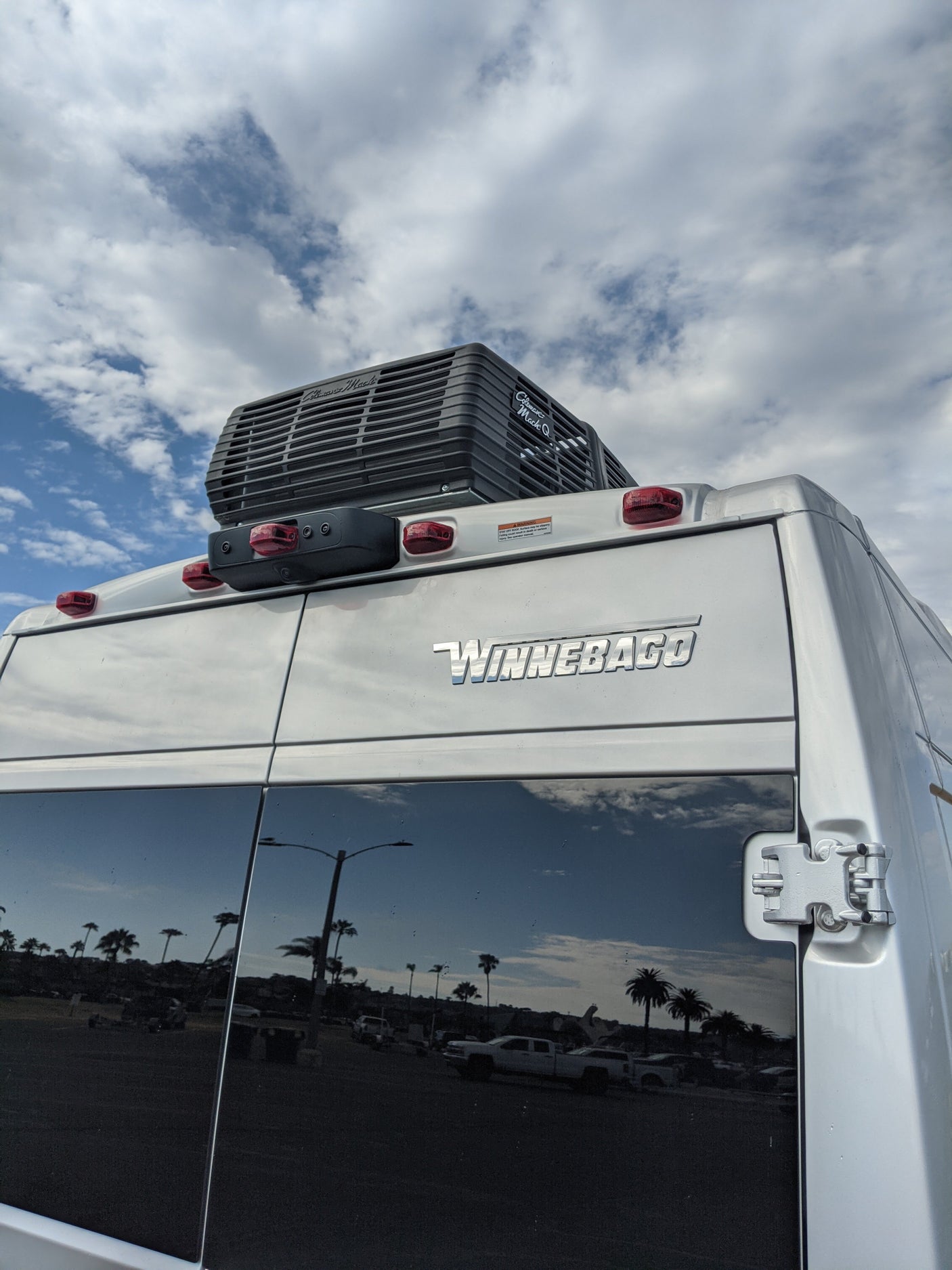 Rear view of a Winnebago RV rental showing its rooftop AC, tail lights, and reflections of palm trees and sky in its tinted windows.