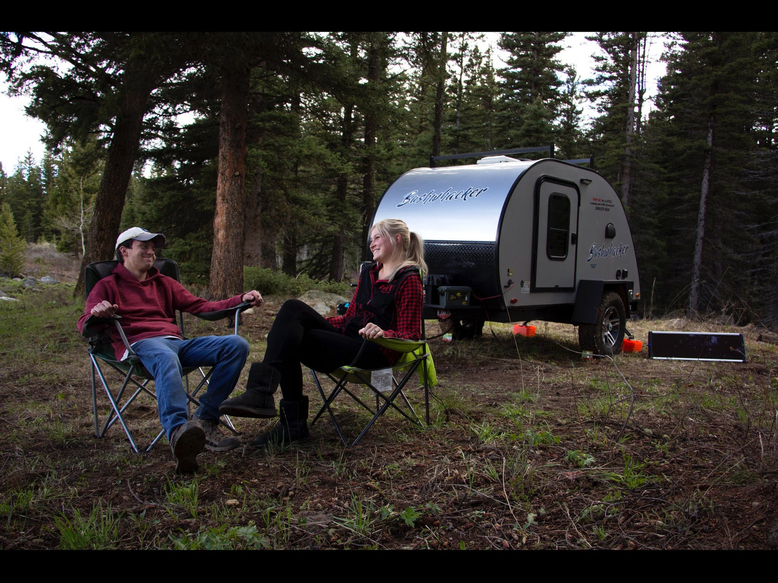 Two people sit in chairs by a Bushwhacker RV rental at a wooded campsite, enjoying the outdoors.