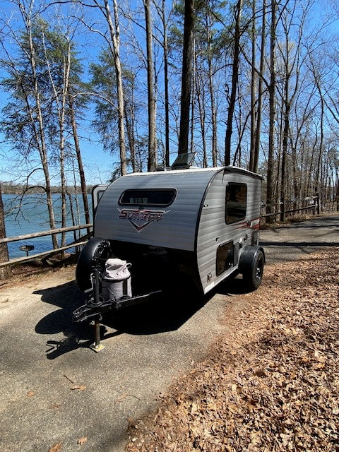 A compact RV rental is set up near a tranquil lake and trees, perfect for a peaceful camping getaway.