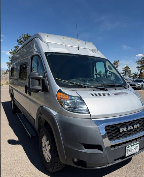 This silver Ram ProMaster rv rental, a camper van with a raised roof, is parked on asphalt under a blue sky, ready for adventure.