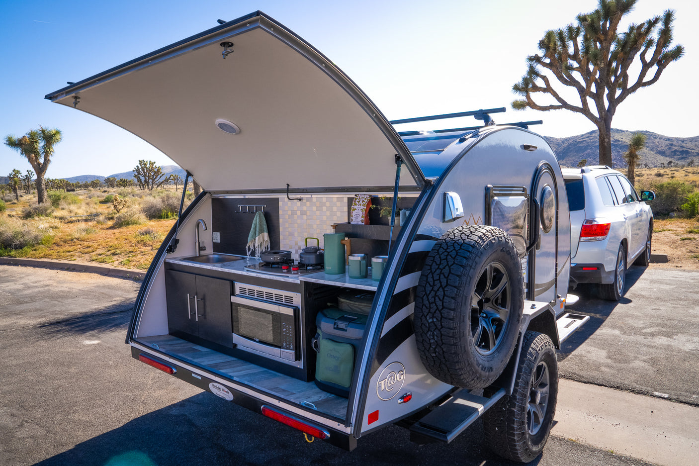 This RV rental teardrop camper, revealing its outdoor kitchen, is set up in a Joshua Tree landscape with an SUV.